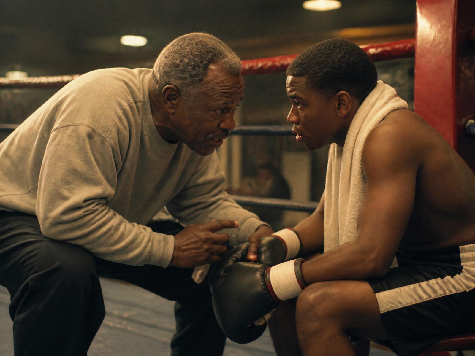 A boxing coach leans in close, speaking quietly to a seated fighter between rounds in a neighborhood gym.